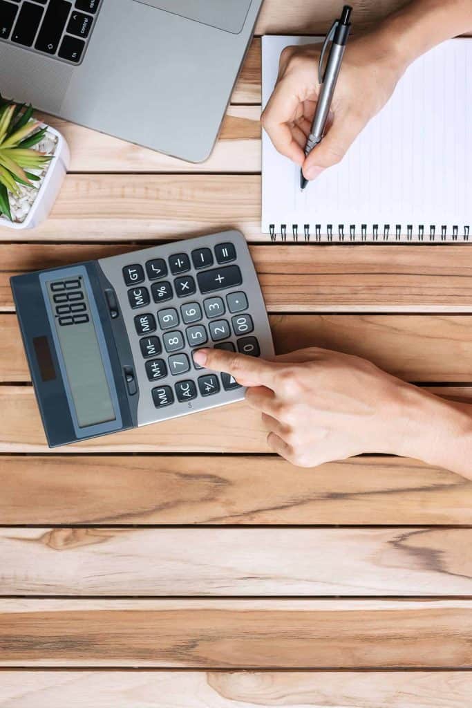 Top view Businesswoman hand writing and calculating with calculator, pen, computer laptop and plant pot on wood office desk table background. workspace or home office with copy space for text concept
