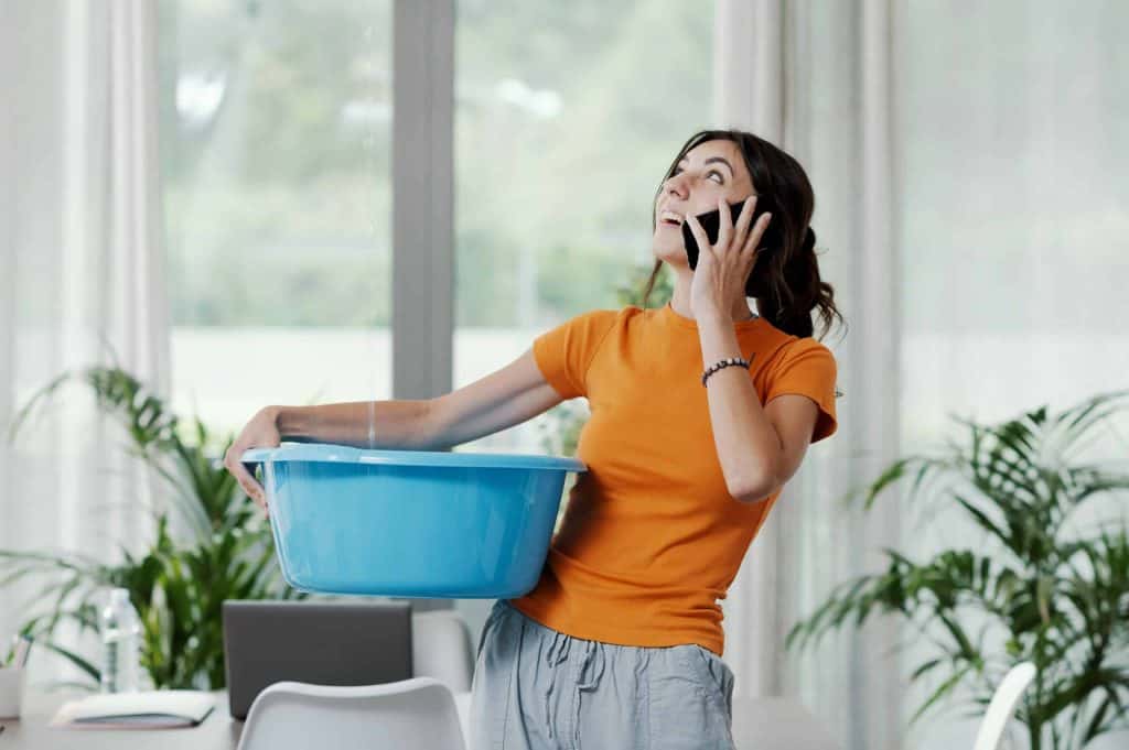 Young upset woman collecting water leaking and dripping from the ceiling, she is calling the roofer